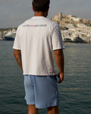 A man stands with his back to the camera by the water, wearing a white t-shirt with 'I AVOID TAX NOT AVADE' printed across the upper back in red and gray, and light blue vertically striped shorts.