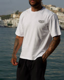 A man wearing a white short-sleeve crewneck t-shirt with a black 'VIESTD WORLD' logo on the left chest, paired with black pleated shorts, standing outdoors with a coastal city and water in the background.