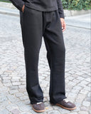 A person wearing black wide-leg trousers, a black long-sleeve top, and brown boat shoes, standing on a cobblestone street with green foliage in the background.