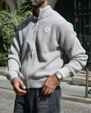 A man wearing a light gray waffle-knit zip-up sweater and dark gray jeans, standing on a cobblestone street in front of a historic building.