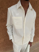 A man wearing a white long-sleeve button-up shirt with a chest pocket and matching white trousers, standing against a textured brown wall.