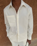 A man wearing a white long-sleeve button-down shirt with a chest pocket and matching white trousers, standing against a textured brown wall.
