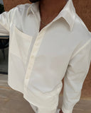 A man wearing a white long-sleeve button-up shirt with a chest pocket, photographed from the neck down against a textured brown wall.