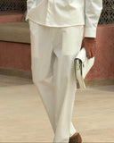 A man wearing a white long-sleeve button-up shirt and matching white trousers, holding a white clutch bag, standing in front of a decorative screen.