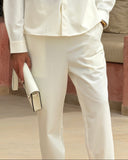 A man wearing a matching off-white two-piece suit with a long-sleeve button-up shirt and trousers, holding a white clutch, standing in front of a decorative stone wall.