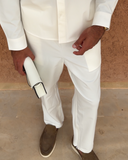 A man wearing a white long-sleeve button-up shirt and matching white trousers, standing against a textured brown wall and holding a white cylindrical bottle.